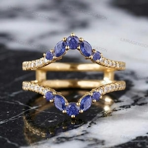 Könnte beinhalten: Ein goldener Ring-Guard bestehend aus zwei V-förmigen Bändern, die durch vertikale Stege verbunden sind. Die Bänder sind mit kleinen, runden, klaren Steinen besetzt und mit blauen Steinen im Marquise-Schliff in V-Form verziert.