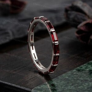 Puede incluir: Un anillo plateado con piedras preciosas rectangulares de color rojo intenso engastadas alrededor de la banda. El anillo se muestra sobre una superficie de madera oscura, con las piedras preciosas captando la luz. Es una joya.