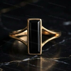 Könnte beinhalten: Ein goldener Ring mit einem vertikalen, rechteckigen schwarzen Edelstein in einer glatten Metallfassung. Die Ringschiene teilt sich in zwei dünne, symmetrische Arme, die zur zentralen Fassung führen und ein klares, geometrisches Design bilden.