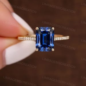 Könnte beinhalten: Ein goldener Ring mit einem großen, rechteckigen, tiefblauen Edelstein. Das goldene Band ist mit kleinen, klaren Steinen besetzt. Der Ring wird vor einem unscharfen Hintergrund präsentiert.
