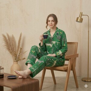 Op de afbeelding: Een groene pyjamaset met lange mouwen en een wit-zwart dierenprintpatroon van luipaarden en zebra's met tropisch gebladerte. Het overhemd met knoopsluiting heeft een reverskraag en witte biezen, passend bij de bijbehorende rechte pyjamabroek.