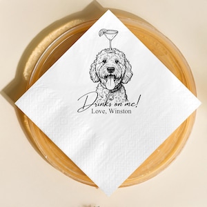 May include: White square cocktail napkin with a black and white illustration of a dog with a martini glass on its head. The text "Drinks on me! Love, Winston" is written below the dog.