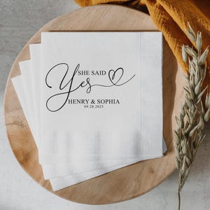 May include: White paper napkin with black calligraphy that says "She Said Yes" with a heart and the names "Henry & Sophia" and the date "09.28.2025".