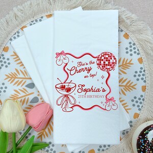 May include: White paper napkins with a red cherry and disco ball design. The text reads "She's the Cherry on top! Sophia's 25th Birthday". The napkins are folded and stacked, with a floral and decorative background.