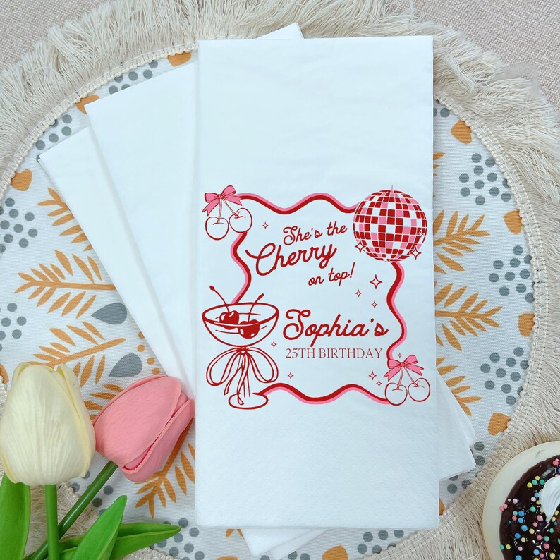 May include: White paper napkins with a red cherry and disco ball design. The text reads "She's the Cherry on top! Sophia's 25th Birthday". The napkins are folded and stacked, with a floral and decorative background.
