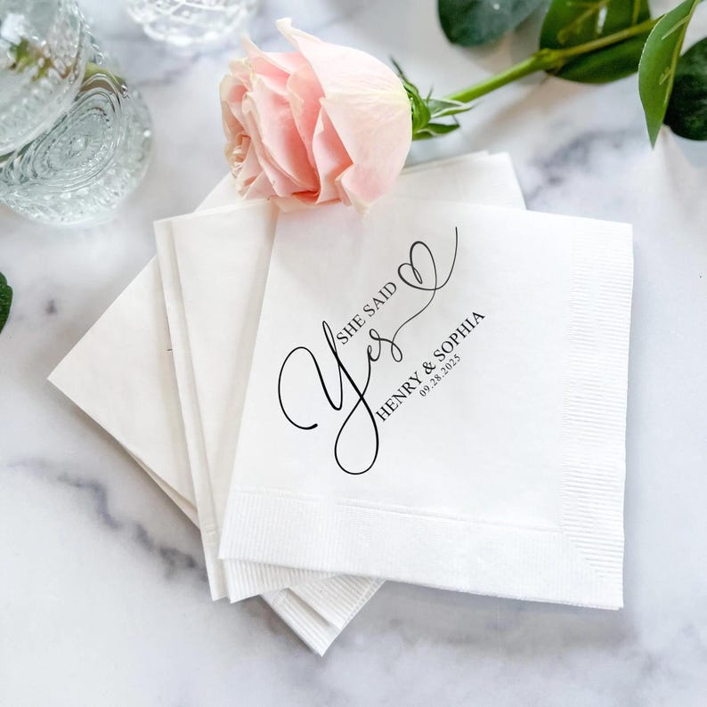 May include: White napkins with black calligraphy that says "She said Yes" with a heart and the text "Henry & Sophia 09.28.2023".