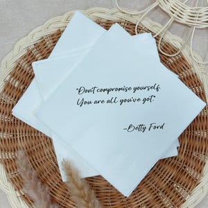 Peut inclure: Serviettes carrées blanches avec la citation "Don't compromise yourself. You are all you've got." de Betty Ford. Les serviettes sont empilées sur un dessous de plat en osier tressé.
