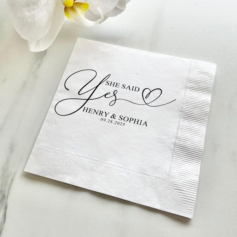 May include: White paper napkin with black calligraphy that says "She Said Yes" with a heart symbol. Below the text is "Henry & Sophia" and the date "09.28.2023".