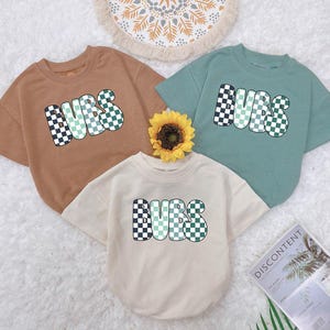 May include: Three t-shirts with the word "BURBS" in checkered green and white letters. The shirts are brown, light green, and cream.