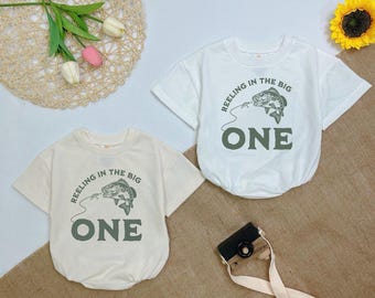 May include: Two cream colored short-sleeved shirts with a green fish graphic and the text "Reeling in the Big One".
