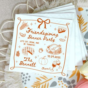 May include: White Friendsgiving dinner party napkins with an orange border and illustrations. The napkins feature text that reads "Friendsgiving Dinner Party" and "Let's eat, drink, and be thankful!" with the date 10.29.2025.