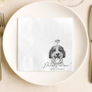 May include: A white paper napkin featuring a black and white line drawing of a dog with its tongue out and a cocktail glass balanced on its head. The text reads, "Drinks on me!" followed by the name "LOVE, TRUMAN" in black ink.