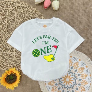 May include: White baby onesie with a green and yellow golf-themed graphic that says "Let's Par-Tee I'm One".