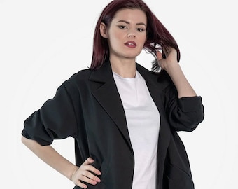 May include: A black blazer with a long, open front and a white t-shirt underneath. The blazer has a collar and two pockets. The person is wearing dark blue jeans.