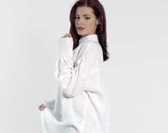 May include: A white linen shirt with long sleeves and a relaxed fit. The shirt has a high neckline and a slightly asymmetrical hem. The model is wearing the shirt with dark blue jeans and white sneakers.