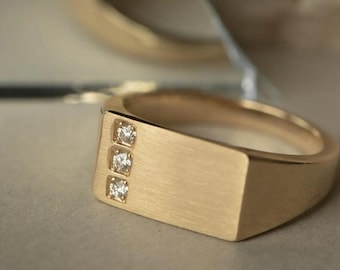 Puede incluir: Anillo dorado con una cara rectangular con tres pequeños diamantes cuadrados. El anillo tiene un acabado de metal cepillado y un diseño sencillo y moderno. El anillo se muestra cerca de un espejo y un fondo texturizado.