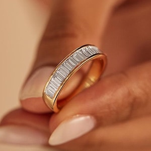 Op de afbeelding: Een gouden ring met een band van rechthoekige, heldere stenen. De ring wordt tussen de vingers gehouden, waarbij het ontwerp en de warme goudkleur worden benadrukt. De ring is een sieraad.