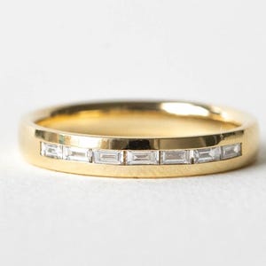May include: A gold band ring with a row of rectangular, clear gemstones. The ring is set against a white background, highlighting the polished gold and the sparkle of the stones. A piece of jewelry.