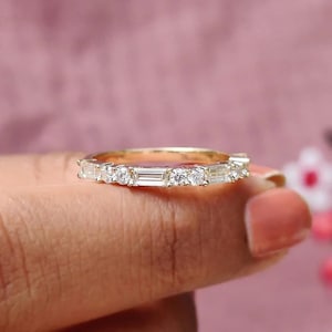 May include: A gold ring featuring a sequence of clear, rectangular and round-cut gemstones. The ring is held by a person, set against a pink backdrop.