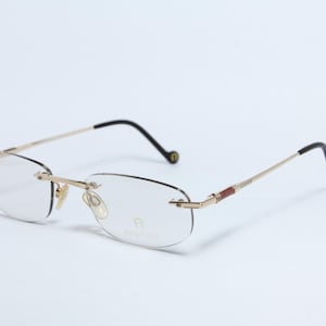 Könnte beinhalten: Randlose Brille mit goldfarbenem Metallrahmen und schwarzen Bügeln. Die Gläser sind klar, und der Markenname "AIGNER" ist auf dem Glas sichtbar. Die Bügel haben einen braunen, lederartigen Akzent.