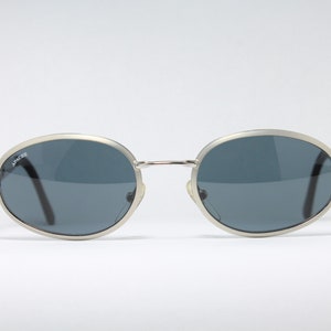 May include: Silver oval-shaped sunglasses with dark gray lenses. The brand name "Apache" is printed on the left lens.