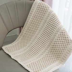 Pode incluir: Uma manta de crochê de cor creme com um padrão de diamantes está drapeada sobre uma cadeira cinza claro.