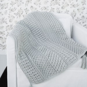May include: A light gray crocheted throw blanket with a textured pattern and tassels on the ends. The blanket is draped over a white armchair.
