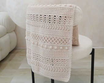 May include: A beige crocheted throw blanket with a delicate pattern and bobble accents. The blanket is draped over a white chair with black legs.
