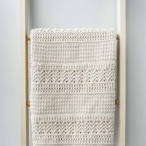 Könnte beinhalten: Eine cremefarbene gehäkelte Wolldecke mit geometrischem Muster, die an einer Holzleiter hängt.