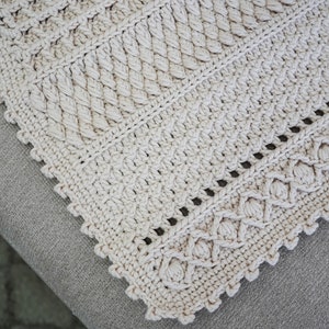 Peut inclure: Gros plan sur une couverture au crochet de couleur beige avec un bord festonn&eacute;. La couverture pr&eacute;sente un motif textur&eacute; avec un motif r&eacute;p&eacute;titif.