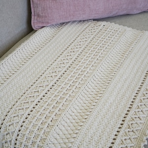 Peut inclure: Une couverture en crochet de couleur cr&egrave;me avec un motif textur&eacute;. La couverture est pli&eacute;e et plac&eacute;e sur un canap&eacute; gris clair.