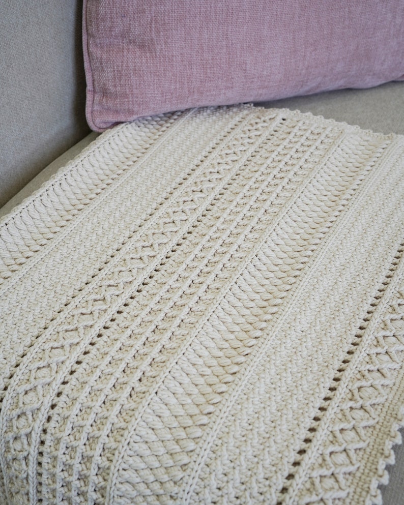 Peut inclure: Une couverture en crochet de couleur cr&egrave;me avec un motif textur&eacute;. La couverture est pli&eacute;e et plac&eacute;e sur un canap&eacute; gris clair.