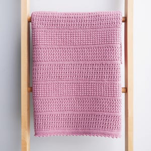 May include: A dusty rose crochet blanket with horizontal textured stripes. The blanket is draped over a light wood ladder. The crochet pattern features a combination of solid and textured stitch work, creating a visually appealing and cozy design.