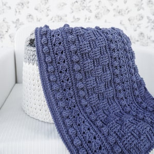 May include: A blue crocheted blanket with a textured pattern and raised bobbles, draped over a white crocheted basket.