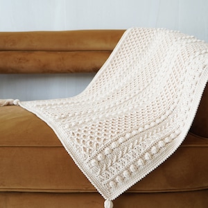 May include: A cream-colored crocheted throw blanket with a textured pattern and tassels on the ends. The blanket is draped over a brown velvet couch.