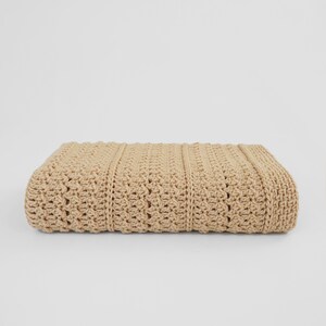 May include: A folded, beige crochet blanket. The blanket features a textured pattern with vertical ribbed sections and a decorative stitch design. The neutral color and detailed craftsmanship suggest a handmade item, suitable for home decor or as a cozy accessory.