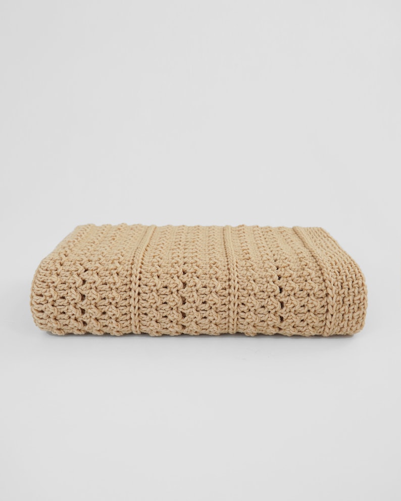 May include: A folded, beige crochet blanket. The blanket features a textured pattern with vertical ribbed sections and a decorative stitch design. The neutral color and detailed craftsmanship suggest a handmade item, suitable for home decor or as a cozy accessory.