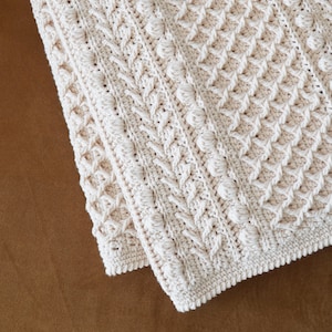 May include: A white crocheted blanket with a textured pattern. The blanket is folded in half and the edges are visible.