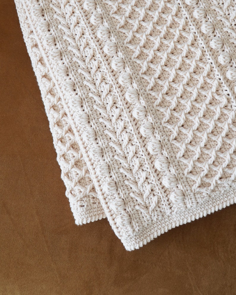 May include: A white crocheted blanket with a textured pattern. The blanket is folded in half and the edges are visible.