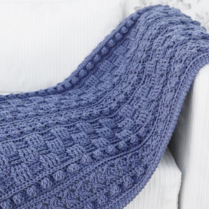 May include: A blue crocheted throw blanket with a textured pattern and bobbles. The blanket is draped over a white armchair.