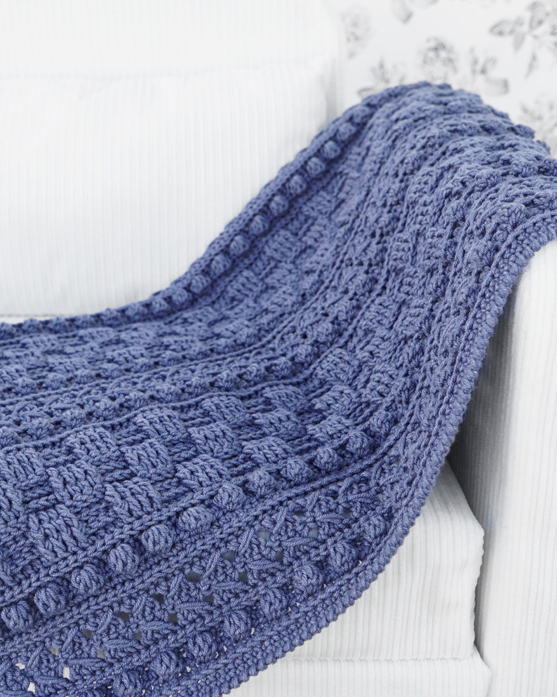 May include: A blue crocheted throw blanket with a textured pattern and bobbles. The blanket is draped over a white armchair.