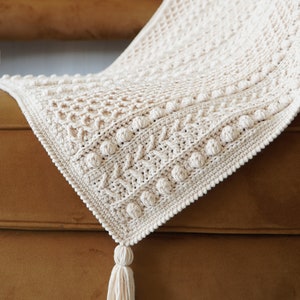 May include: A cream-colored crocheted throw blanket with a textured pattern and a tassel. The blanket is draped over a brown couch.
