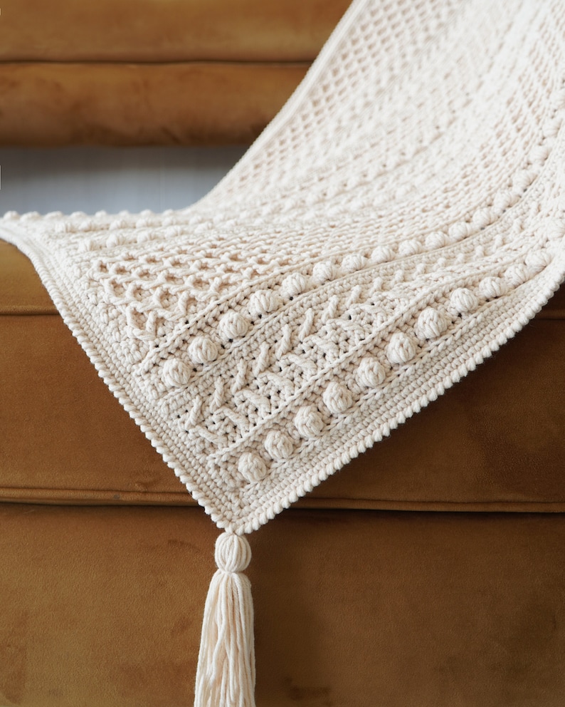May include: A cream-colored crocheted throw blanket with a textured pattern and a tassel. The blanket is draped over a brown couch.