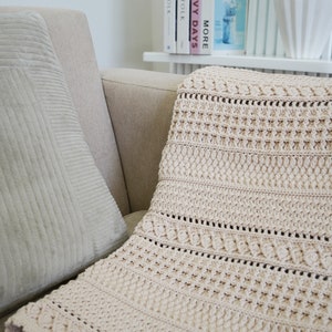 Peut inclure: Une couverture en crochet beige avec un motif textur&eacute;, drap&eacute; sur un canap&eacute; brun clair avec un coussin gris.