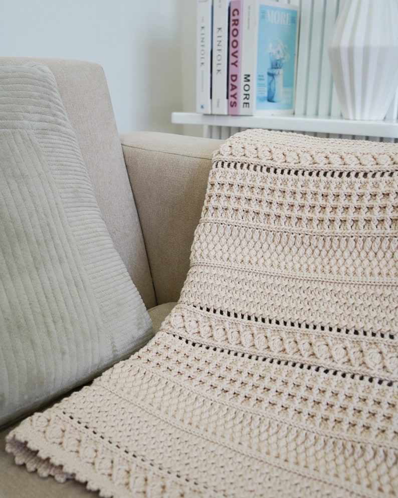 Peut inclure: Une couverture en crochet beige avec un motif textur&eacute;, drap&eacute; sur un canap&eacute; brun clair avec un coussin gris.