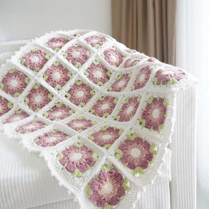 May include: A white crocheted blanket with a floral pattern in shades of pink and green. The blanket is folded and draped over a white sofa.