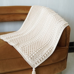May include: A cream-colored crocheted throw blanket with a tassel on a brown velvet couch.
