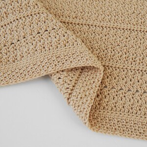 May include: A folded, beige crochet blanket. The blanket has a textured stitch pattern and a finished border. The neutral color and handmade appearance suggest a cozy, home decor item. The blanket is folded over on a white surface.