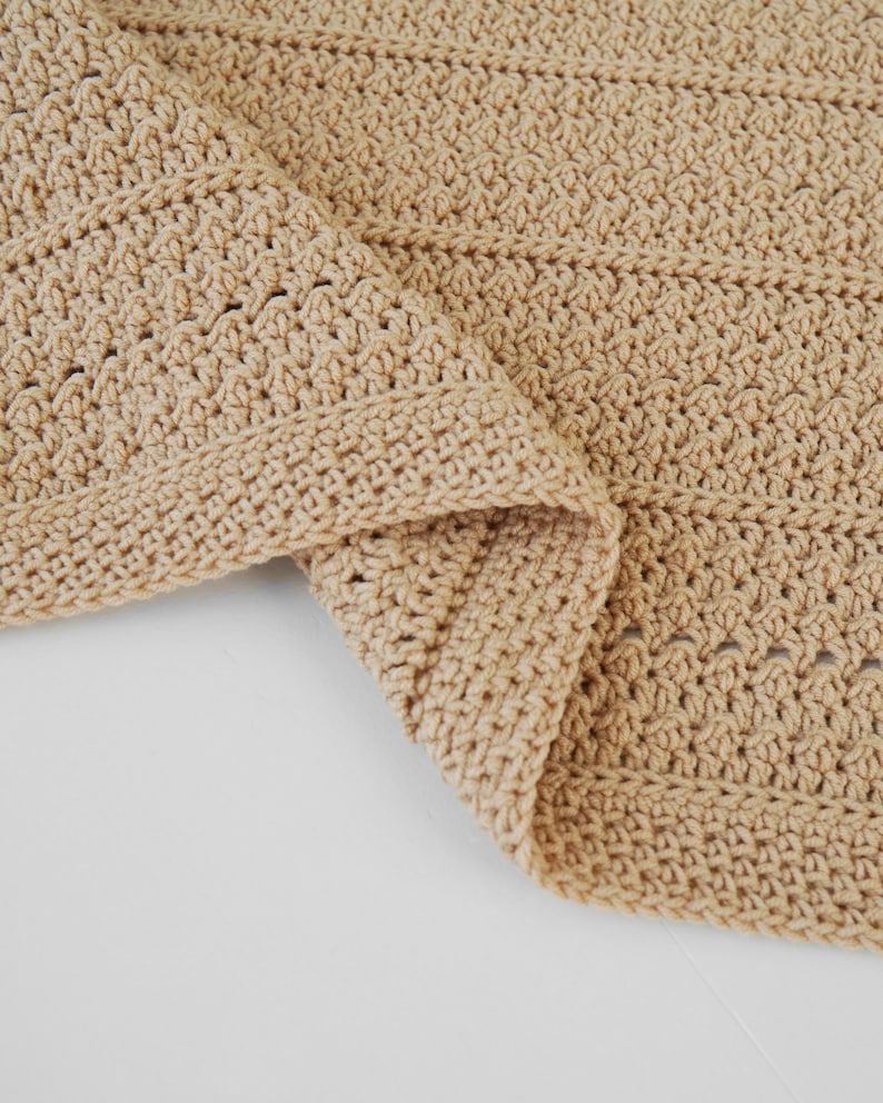 May include: A folded, beige crochet blanket. The blanket has a textured stitch pattern and a finished border. The neutral color and handmade appearance suggest a cozy, home decor item. The blanket is folded over on a white surface.