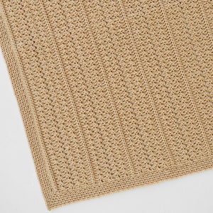 May include: A close-up of a tan crochet blanket. The blanket features a textured pattern with vertical lines and a detailed border. The neutral color and intricate design suggest a handmade item, suitable for home decor or as a cozy accessory.